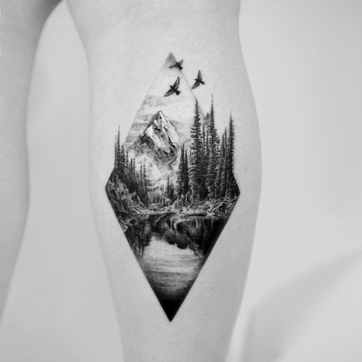 10 Mountain Landscape Tattoo Ideas For Your Next Ink 8 A diamond-shaped black and white tattoo of a mountain, forest, and lake on a person's arm.