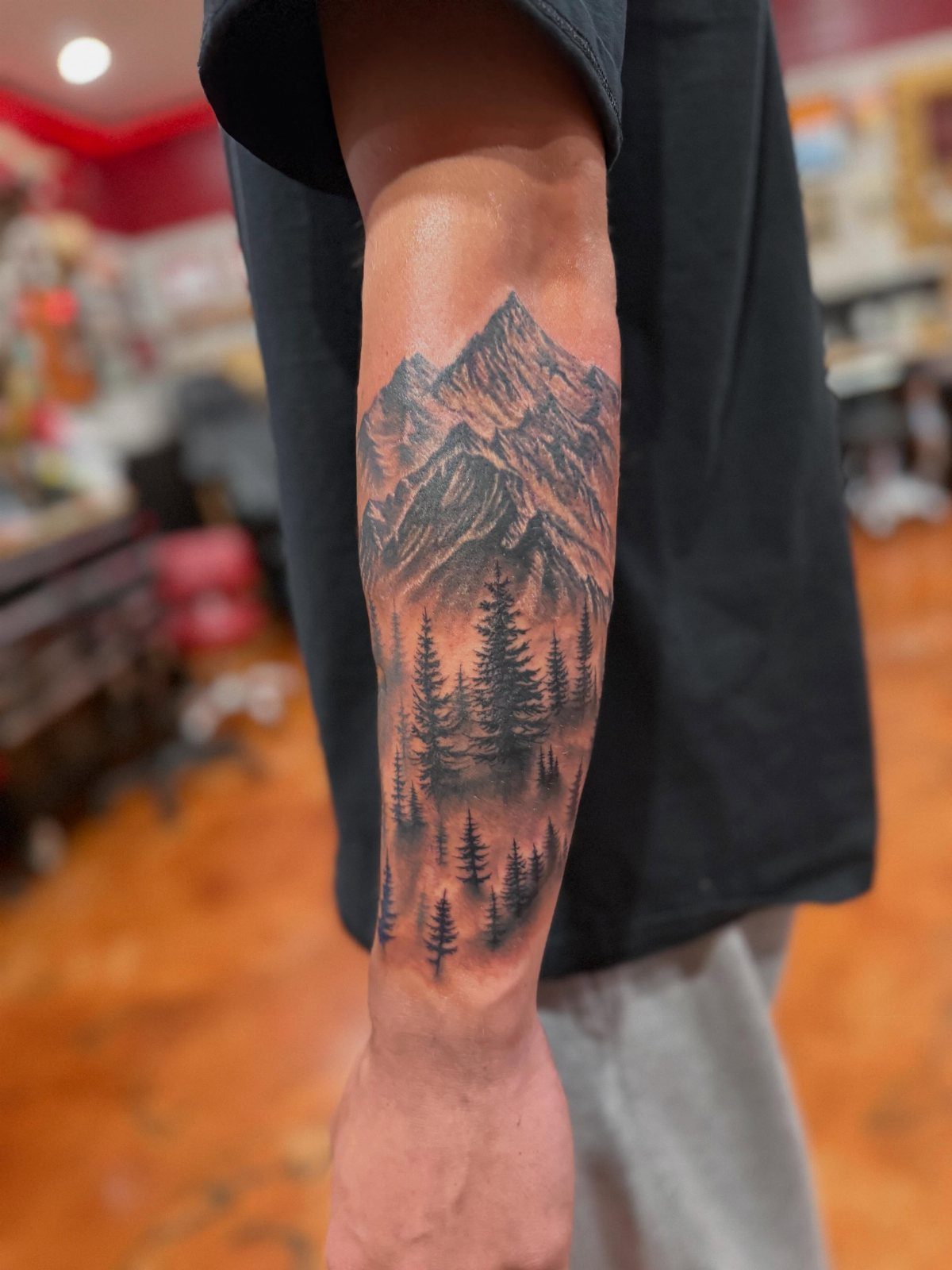 10 Mountain Landscape Tattoo Ideas For Your Next Ink 1 A detailed black and grey mountain landscape tattoo featuring jagged peaks and a pine forest on a forearm.