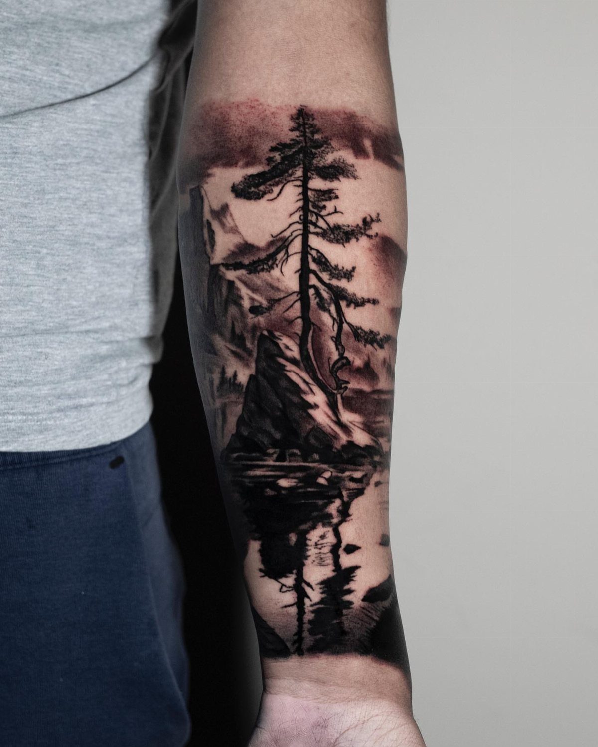 10 Mountain Landscape Tattoo Ideas For Your Next Ink 4 A realistic black and grey forearm tattoo featuring a pine tree, mountains, and a reflective lake.