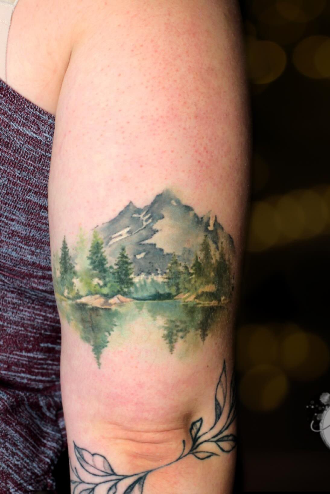 10 Mountain Landscape Tattoo Ideas For Your Next Ink 3 A watercolor mountain landscape tattoo with green trees and a lake on a person's arm.