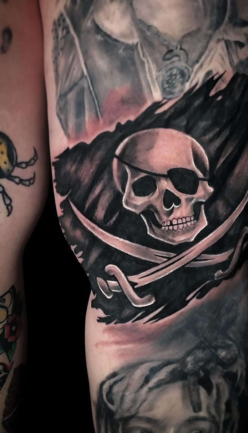 A black and grey tattoo of a skull with an eye patch and crossed swords.