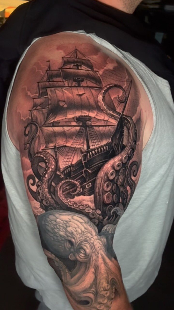 A large black and grey tattoo of a sailing ship being wrapped by octopus tentacles.