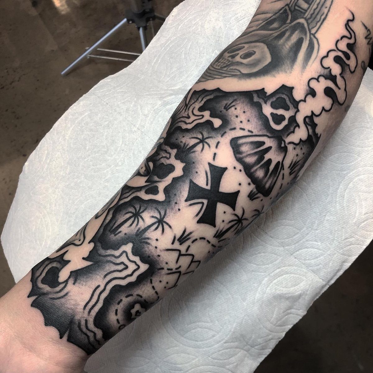A black and grey forearm tattoo featuring a stylized treasure map with coastlines and an X mark.