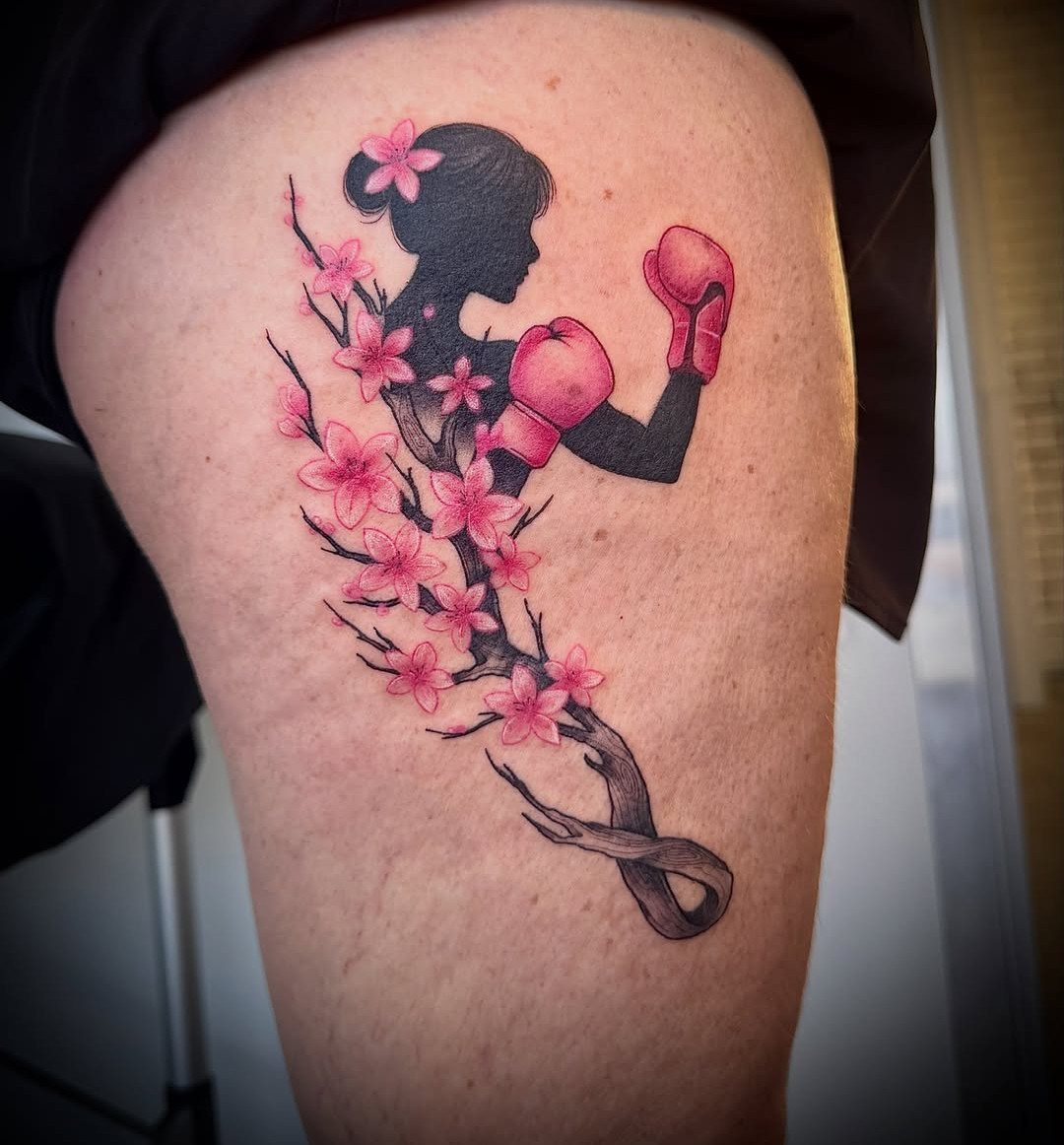 Sickness Tattoo Ideas: 6 Designs That Mean Something 4 A black and pink silhouette tattoo of a female boxer surrounded by cherry blossoms on a person's thigh.