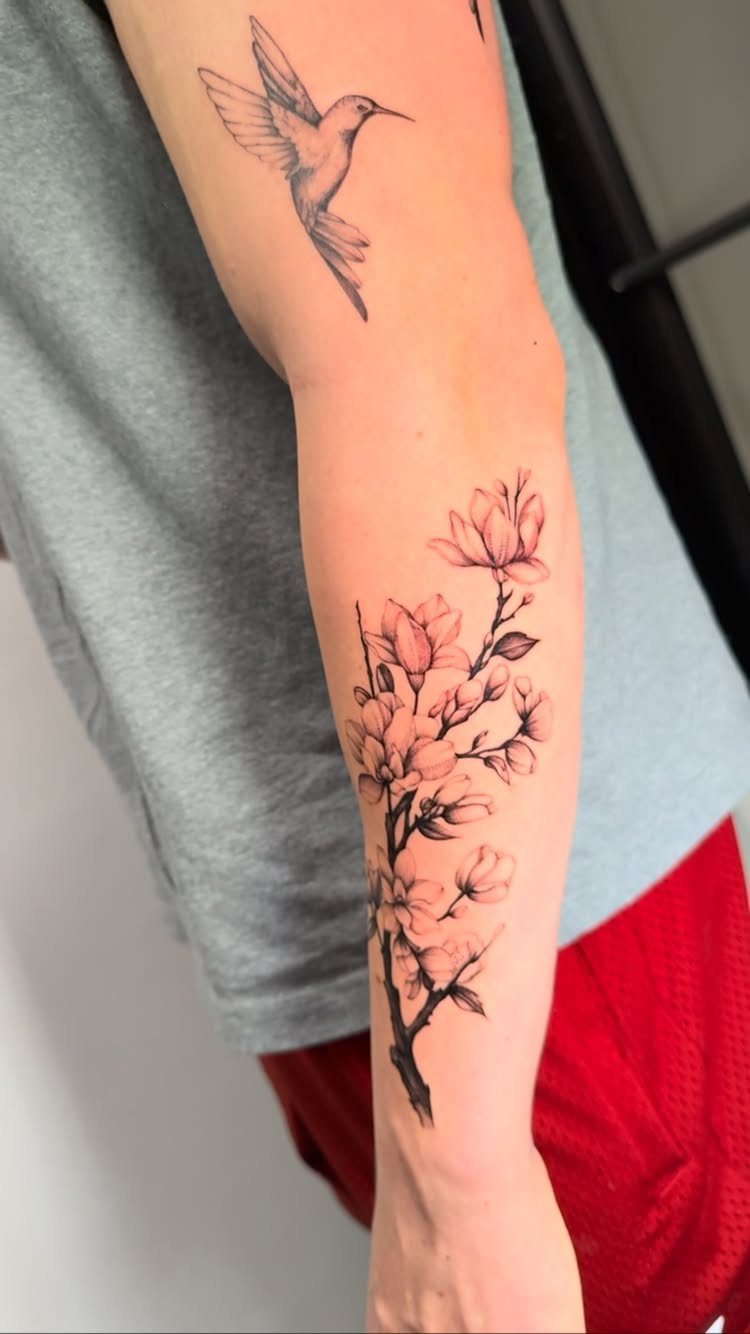 A forearm featuring a detailed black and grey magnolia floral tattoo and a hummingbird tattoo.