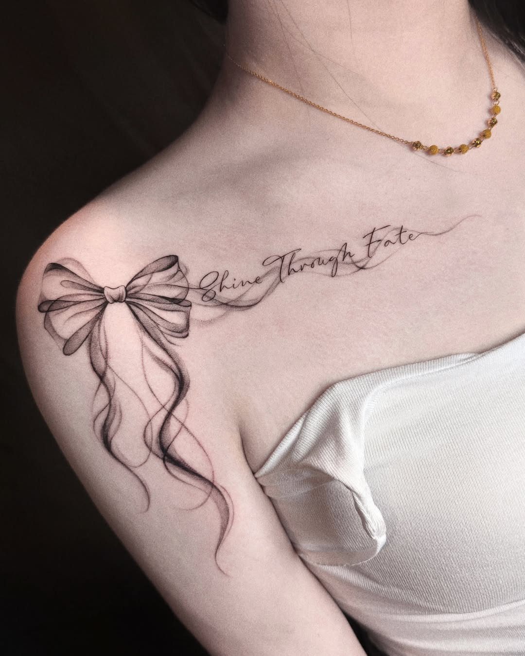 A delicate black ink bow tattoo with script text on a person's collarbone.