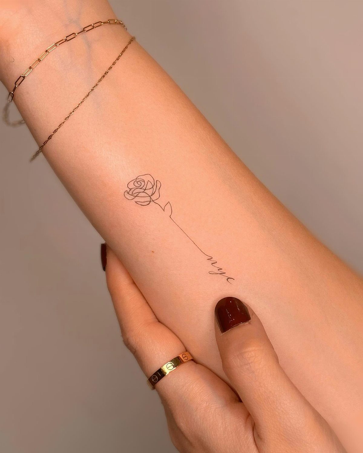 A delicate fine line rose tattoo on a person's forearm with gold jewelry.