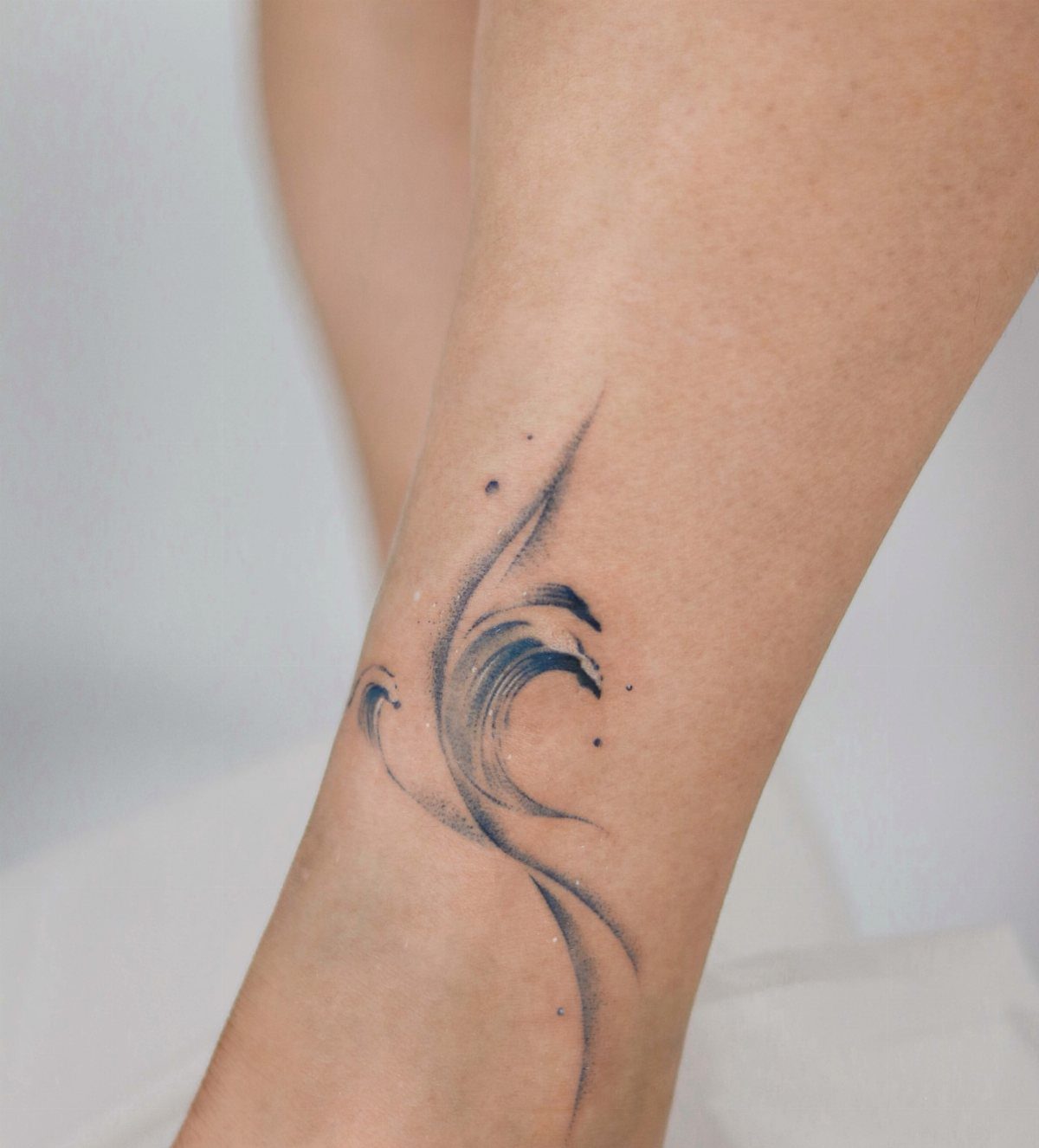 A minimalist blue ink wave tattoo on a person's lower leg skin.