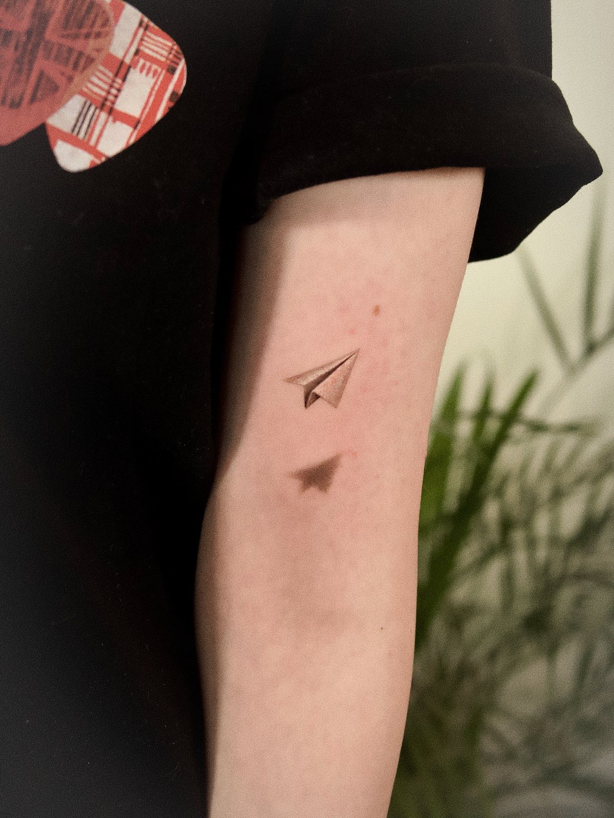 A small black ink paper airplane tattoo on a person's arm with a subtle shadow.