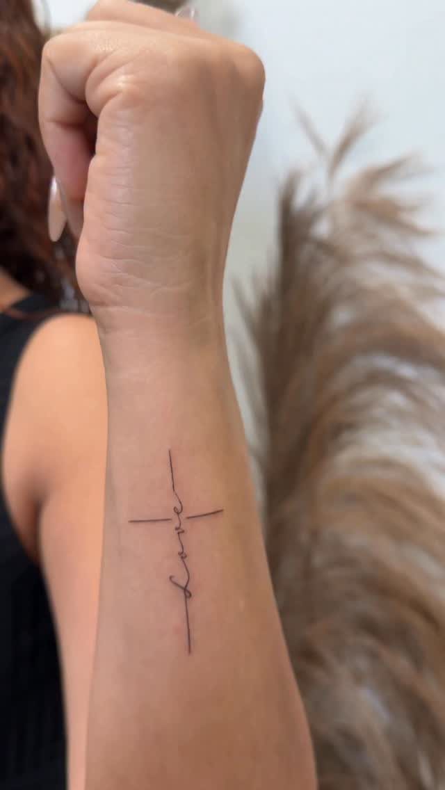 A small black ink tattoo of a cross with the word love written in script on a wrist.