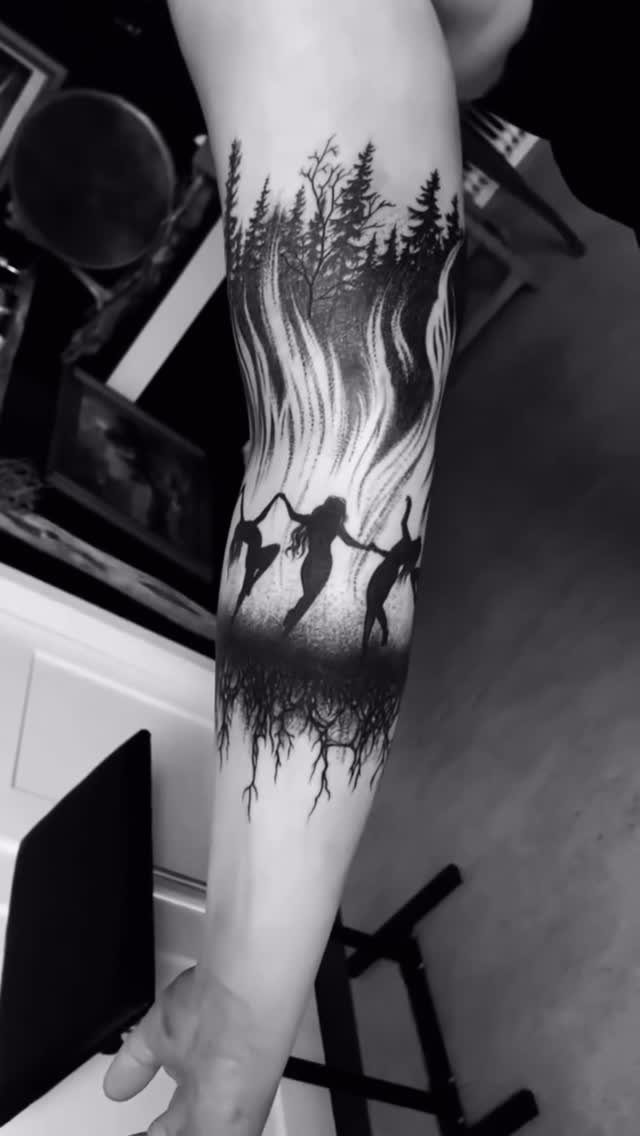 A large black and white leg tattoo depicting a forest, dancing figures, and roots.