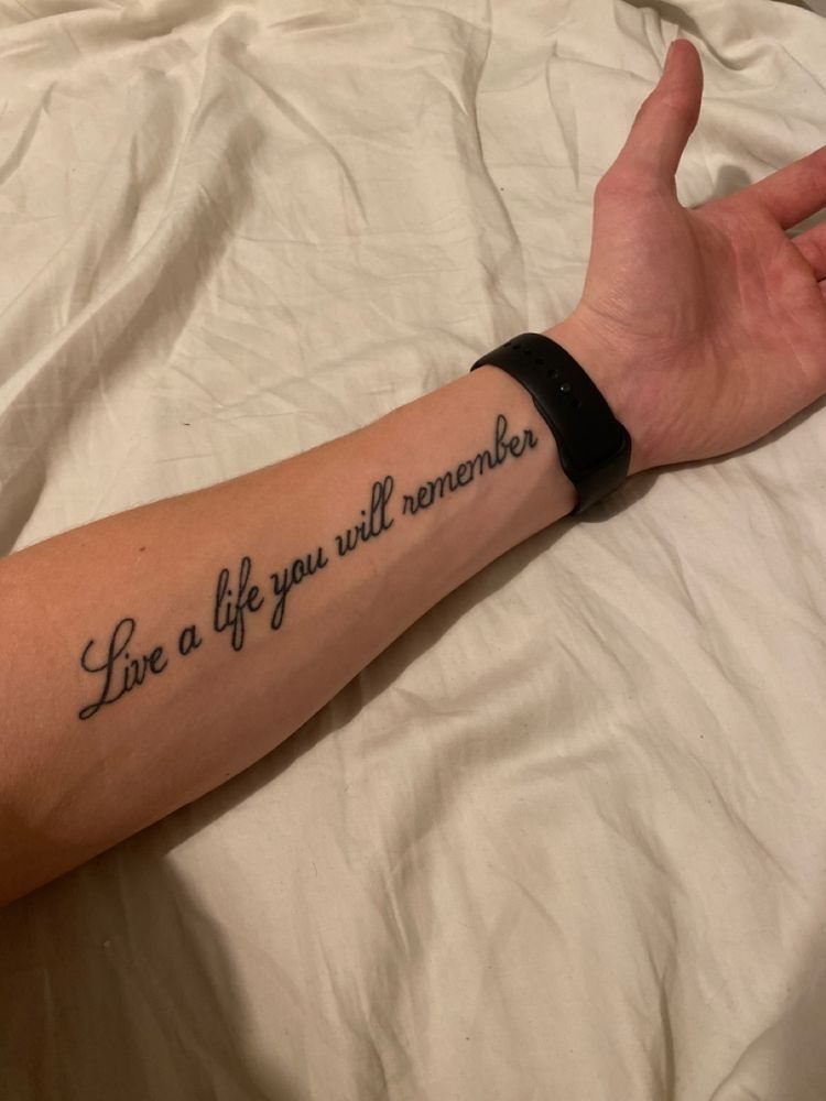 A forearm tattoo in black cursive script reading Live a life you will remember.