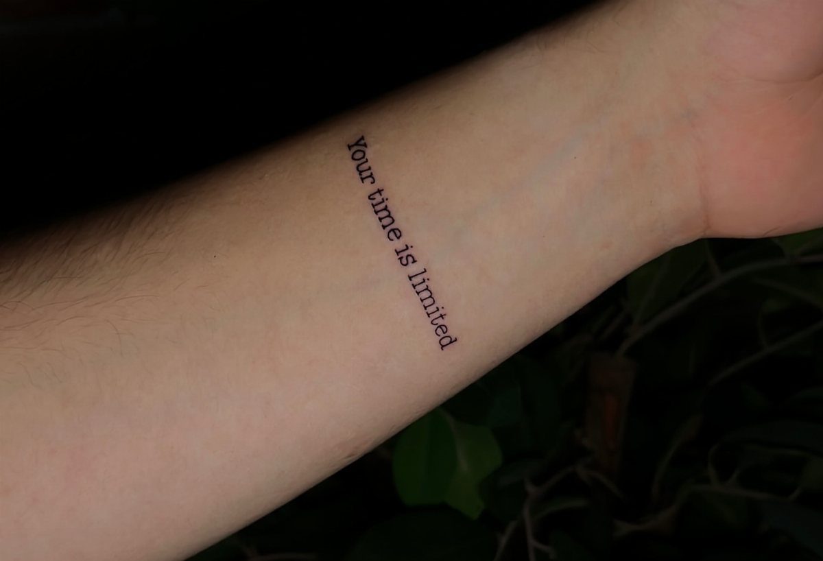 A small black ink text tattoo reading Your time is limited on a person's forearm.