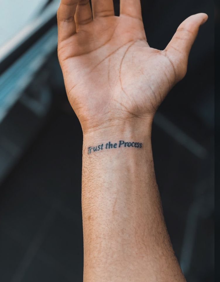 A person's wrist featuring a small black ink tattoo that says Trust the Process.