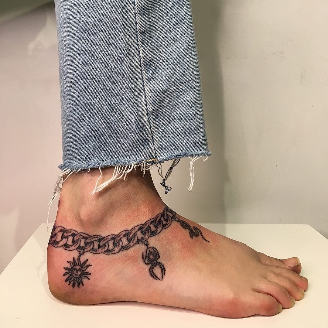 A black ink tattoo of a chain anklet with sun and moon charms on a foot.