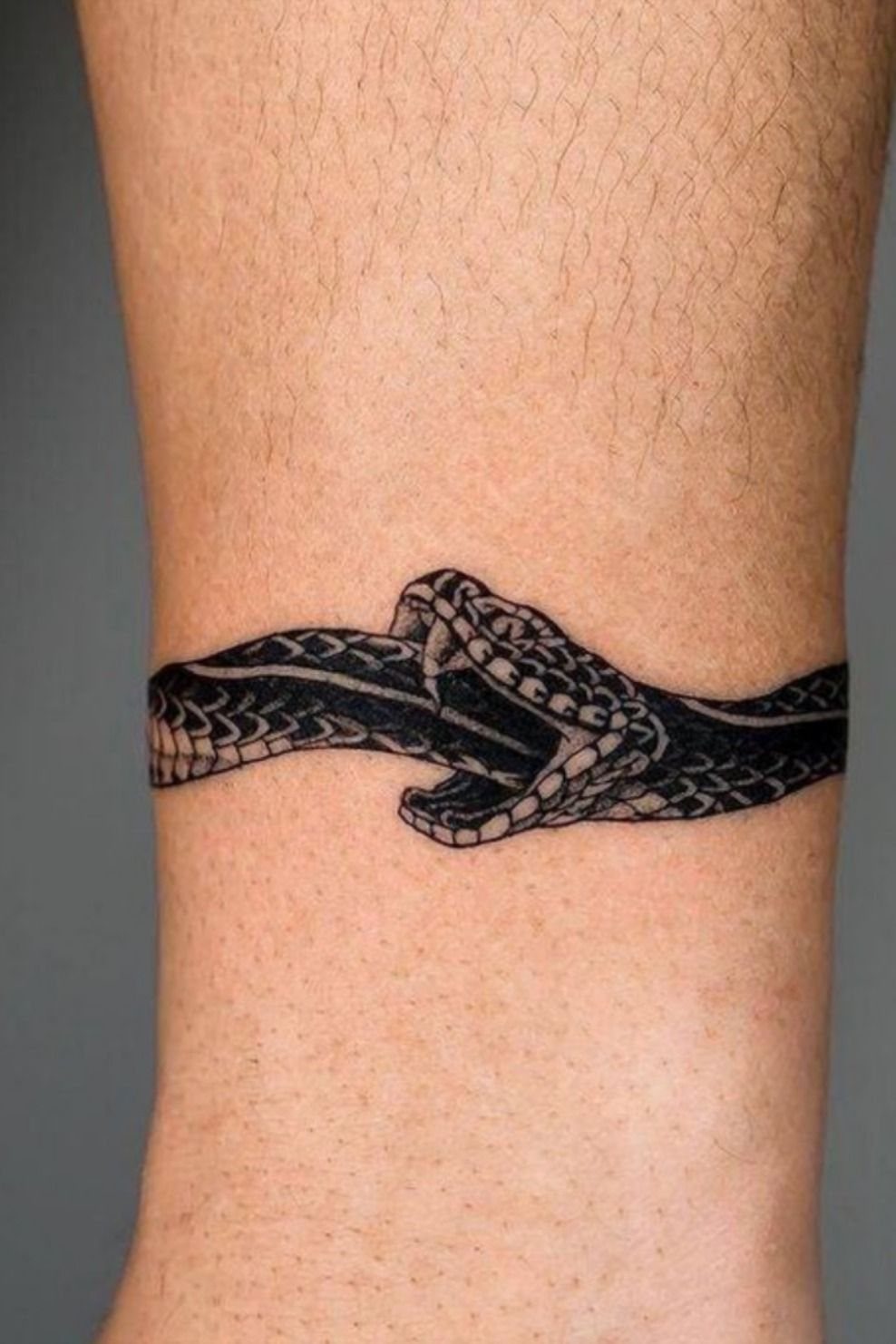 A black ink snake tattoo with an open mouth wrapped around a person's ankle.