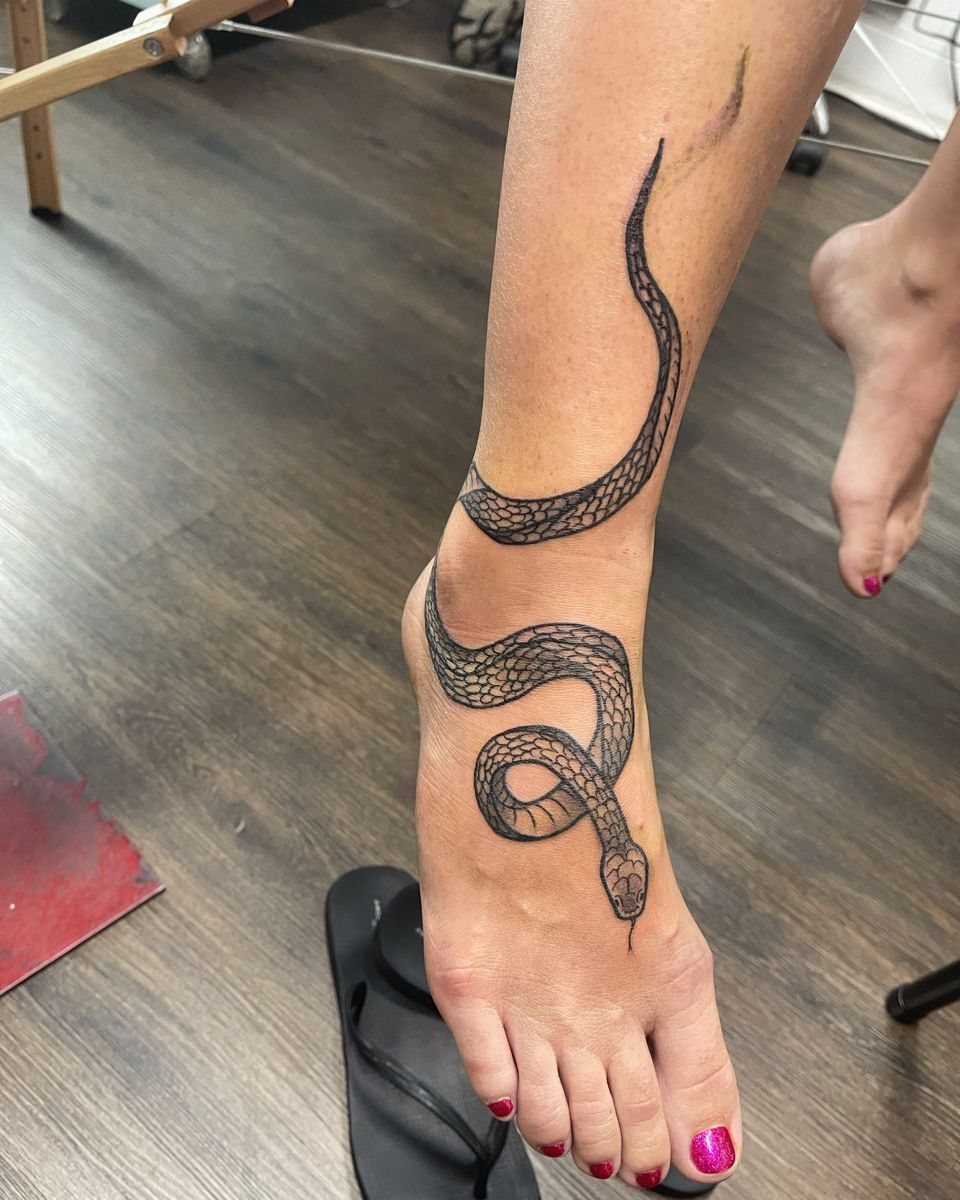 A black ink snake tattoo winding around an ankle and across the top of a foot.