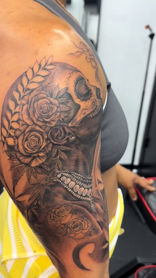 A large black and grey sugar skull tattoo with roses on a person's shoulder.