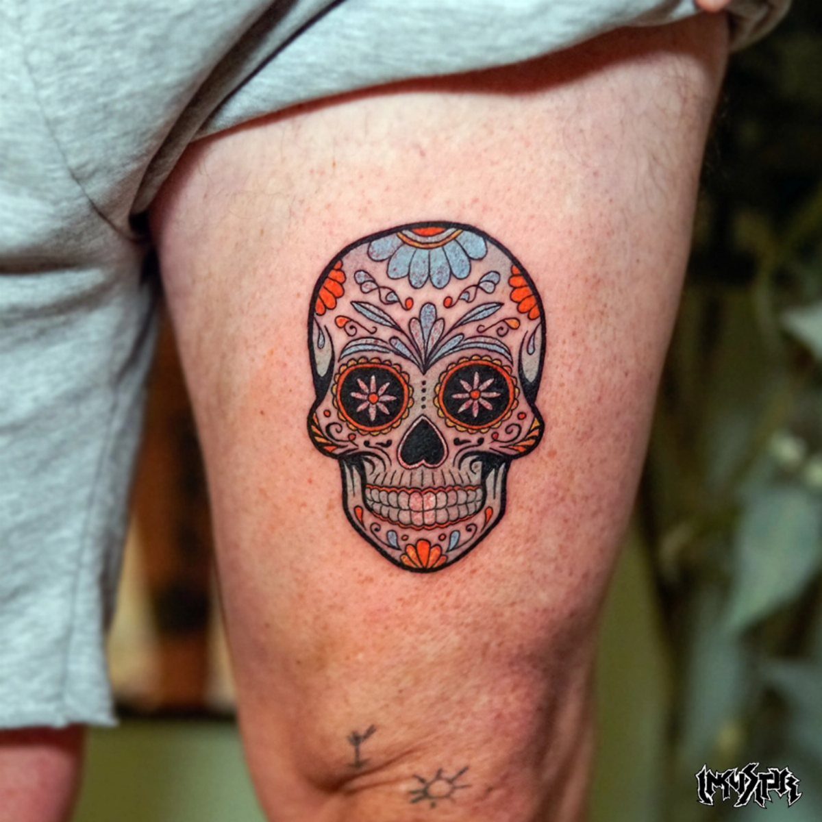 A colorful sugar skull tattoo with orange and blue floral patterns on a person's thigh.