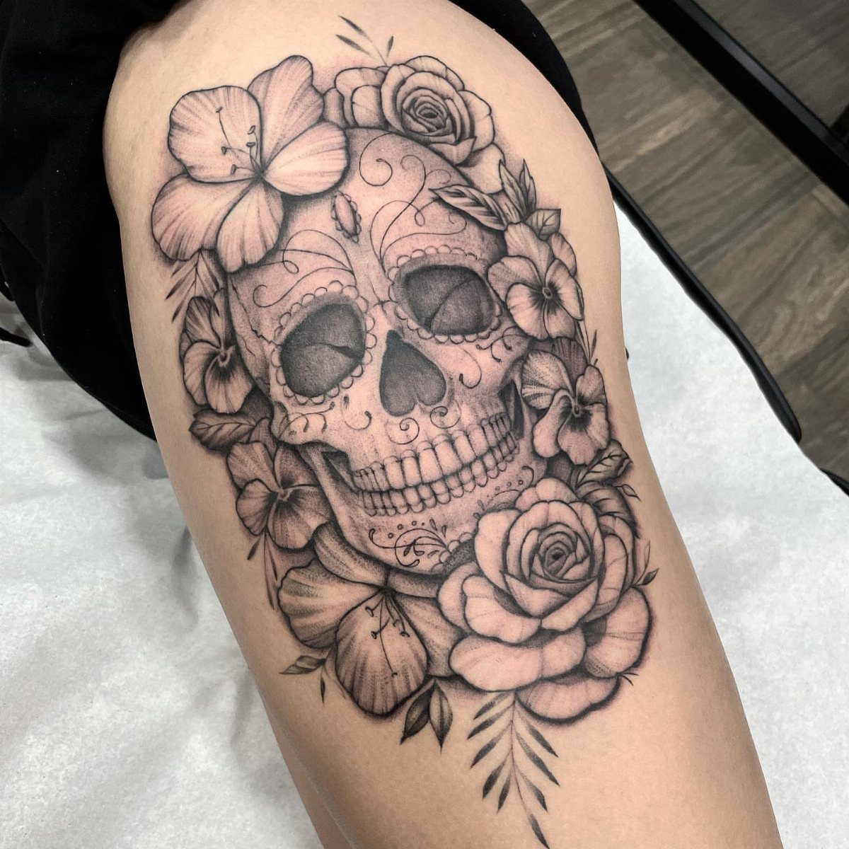 A black and grey sugar skull tattoo decorated with large roses and hibiscus flowers on a leg.