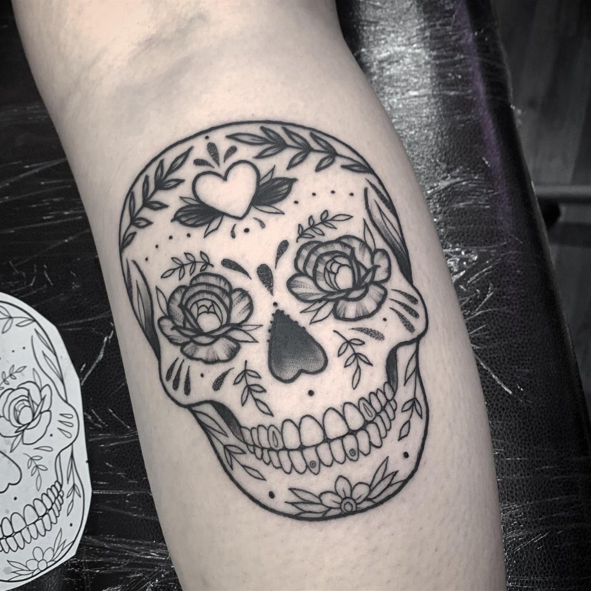 A black and grey sugar skull tattoo with floral patterns and a heart on an arm.