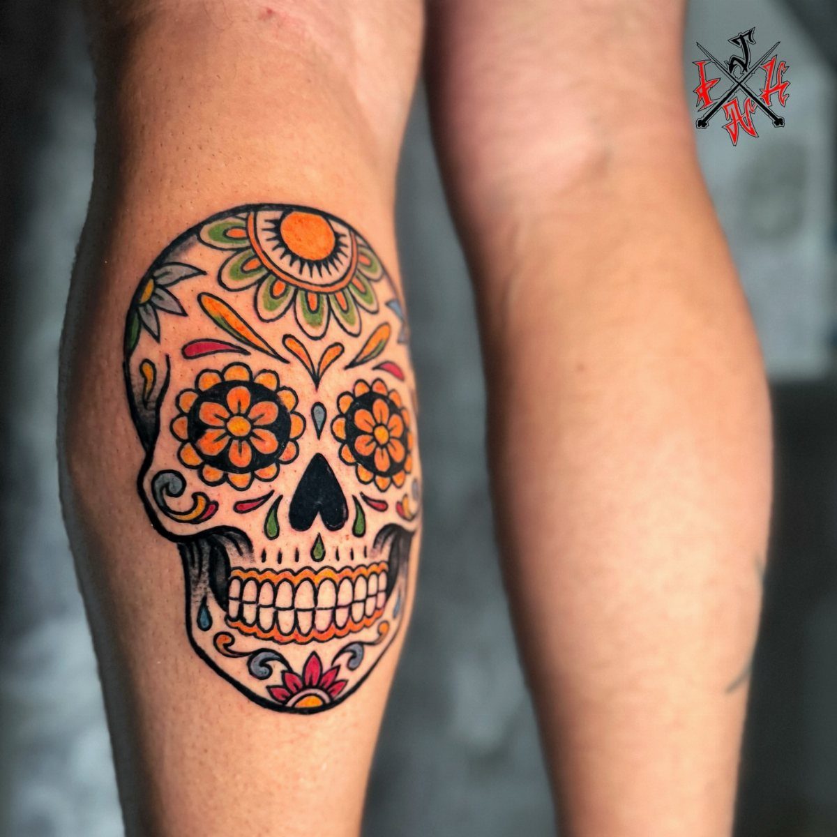 A colorful sugar skull tattoo with orange and yellow floral patterns on a person's leg.