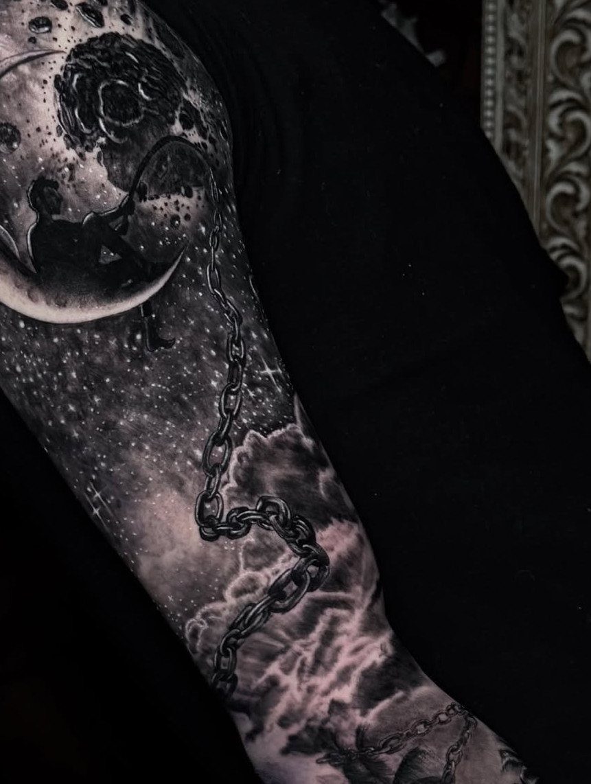 A detailed black and grey tattoo on an arm showing a moon, silhouette, chain, and starry clouds.