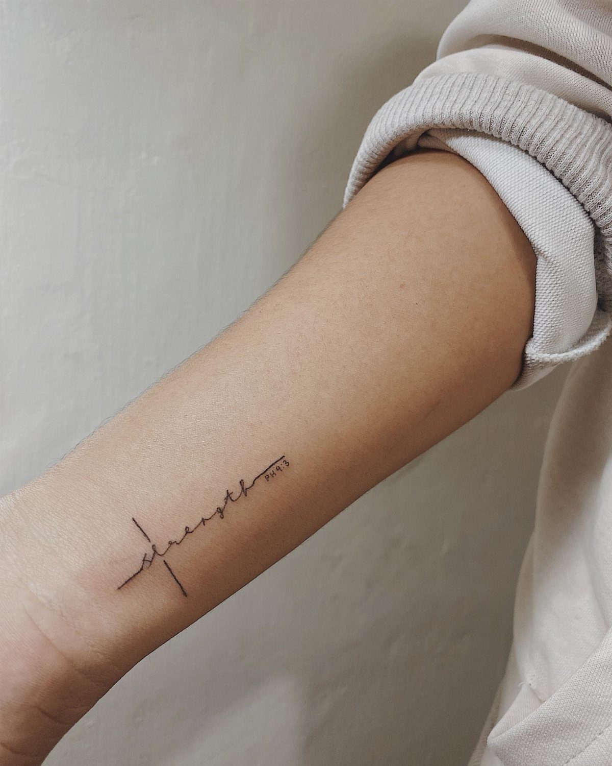 A minimalist black ink tattoo of a cross combined with the word strength on a forearm.