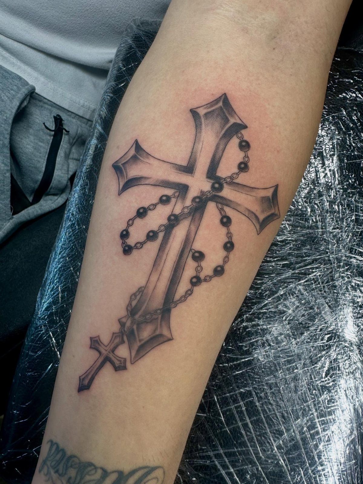 A black and grey tattoo of a stylized cross entwined with a beaded rosary on a forearm.