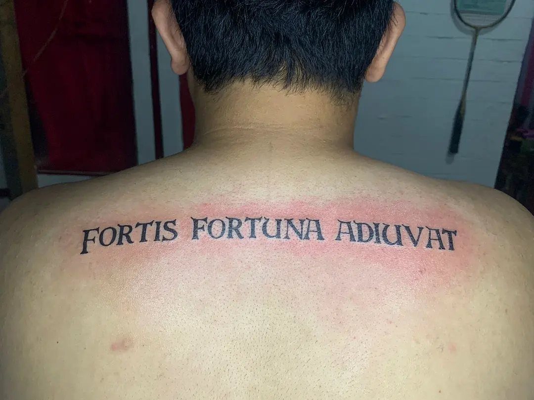 16 Word Tattoo Placement Ideas: From Spine to Finger 6 A person's upper back featuring a black ink Latin phrase tattoo across the shoulder blades.