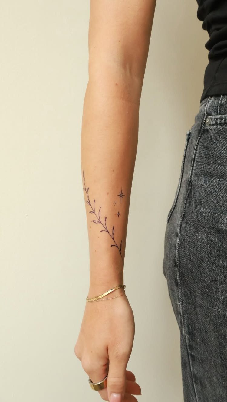 A person's forearm displaying a delicate black line art tattoo of leafy branches and stars.