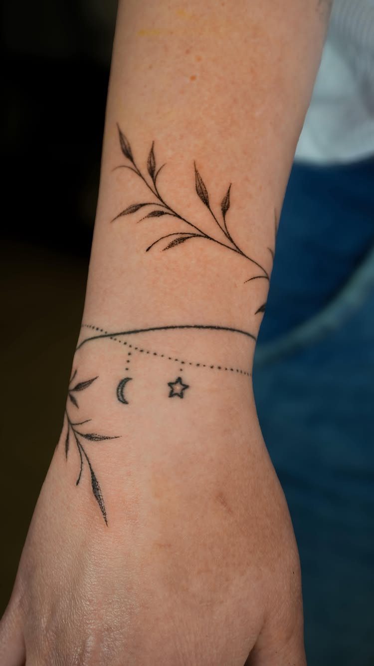 A delicate black ink tattoo of leaves and stars encircling a person's wrist.