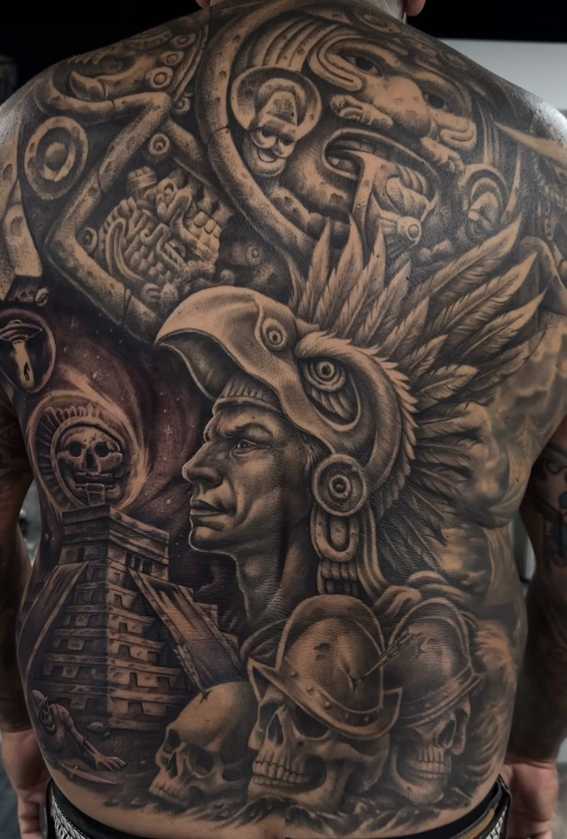 A detailed black and grey back tattoo depicting an Aztec warrior, pyramids, and ancient cultural symbols.