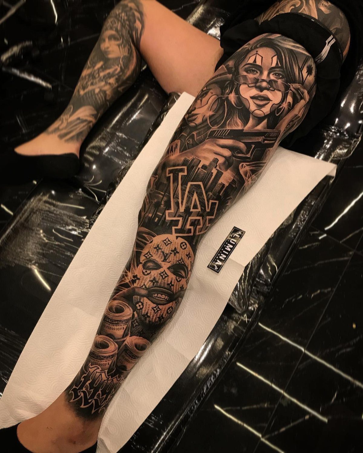 A detailed black and grey leg tattoo featuring a woman's portrait and various Chicano style elements.