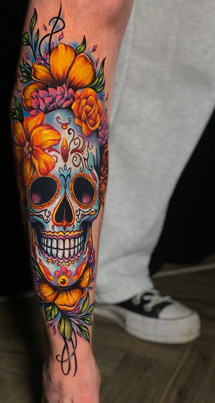 A colorful sugar skull tattoo with bright orange flowers on a person's leg.