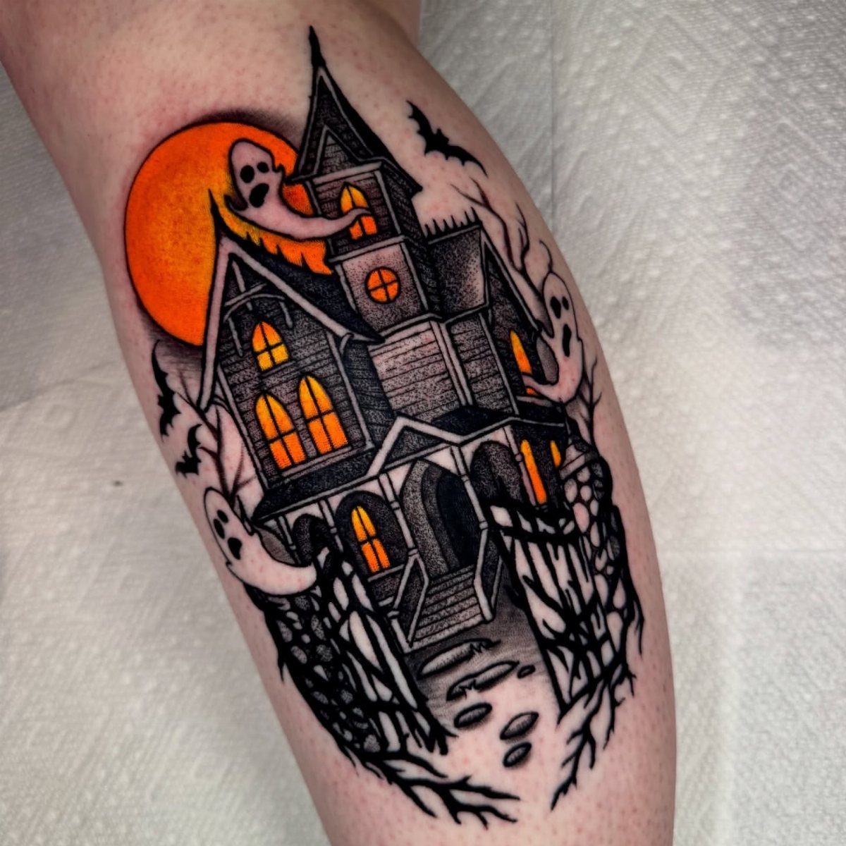 7 Tattoo Style Home Designs for Your Next Ink 1 A spooky haunted house tattoo with orange glowing windows and ghosts on a person's leg.