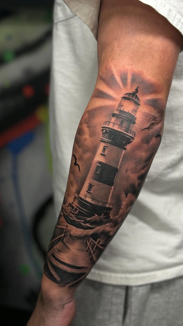 7 Tattoo Style Home Designs for Your Next Ink 2 A black and grey realism tattoo of a lighthouse with light rays on a person's forearm.