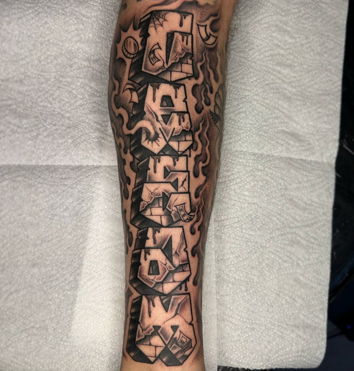 A large black and grey tattoo of geometric block letters on a person's arm.