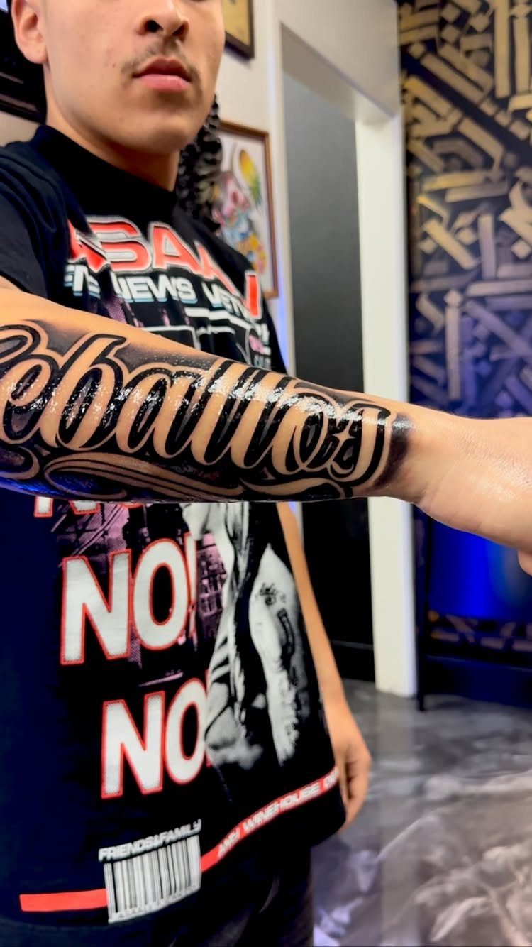 A close up of a person's arm featuring a large Chicano style script tattoo.