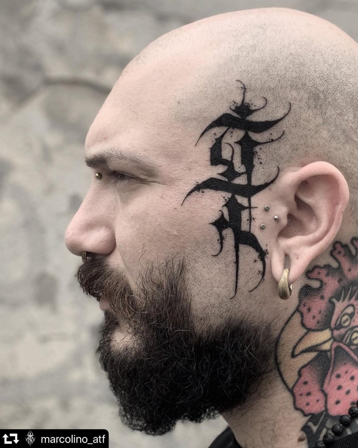 A man with a beard featuring a black gothic script tattoo on his temple and cheek.