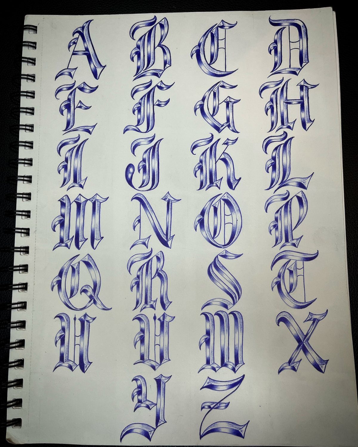 Hand-drawn blue ink alphabet in a decorative gothic tattoo lettering style on notebook paper.