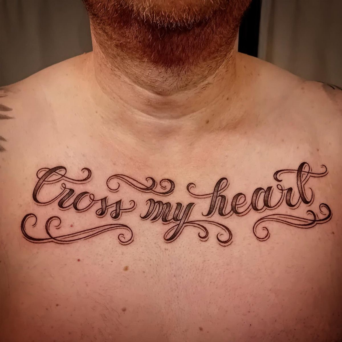 A man's chest featuring a large black ink script tattoo reading Cross my heart.