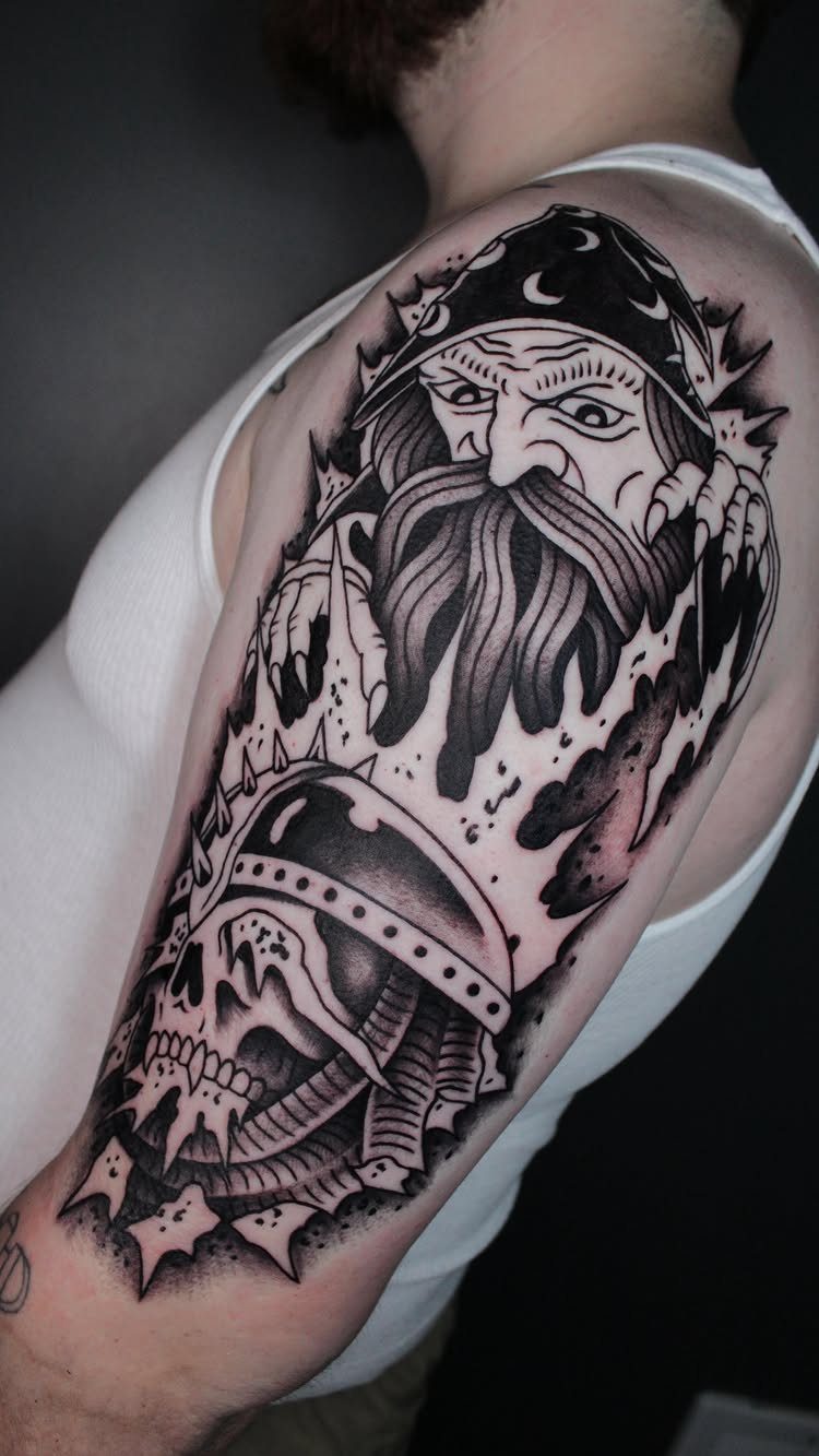 A neo-traditional black and grey tattoo of a bearded man and a skull on a person's arm.