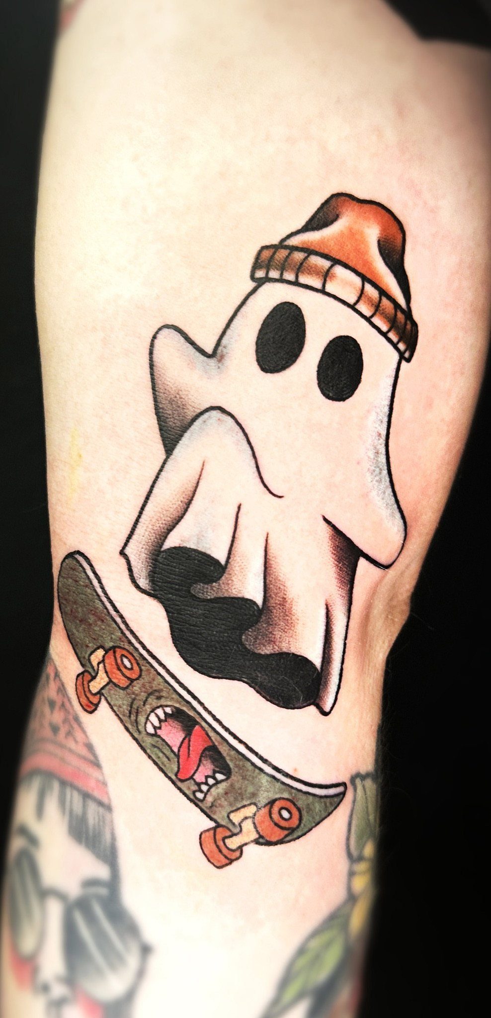 A neo-traditional tattoo of a ghost wearing a beanie and riding a skateboard on skin.