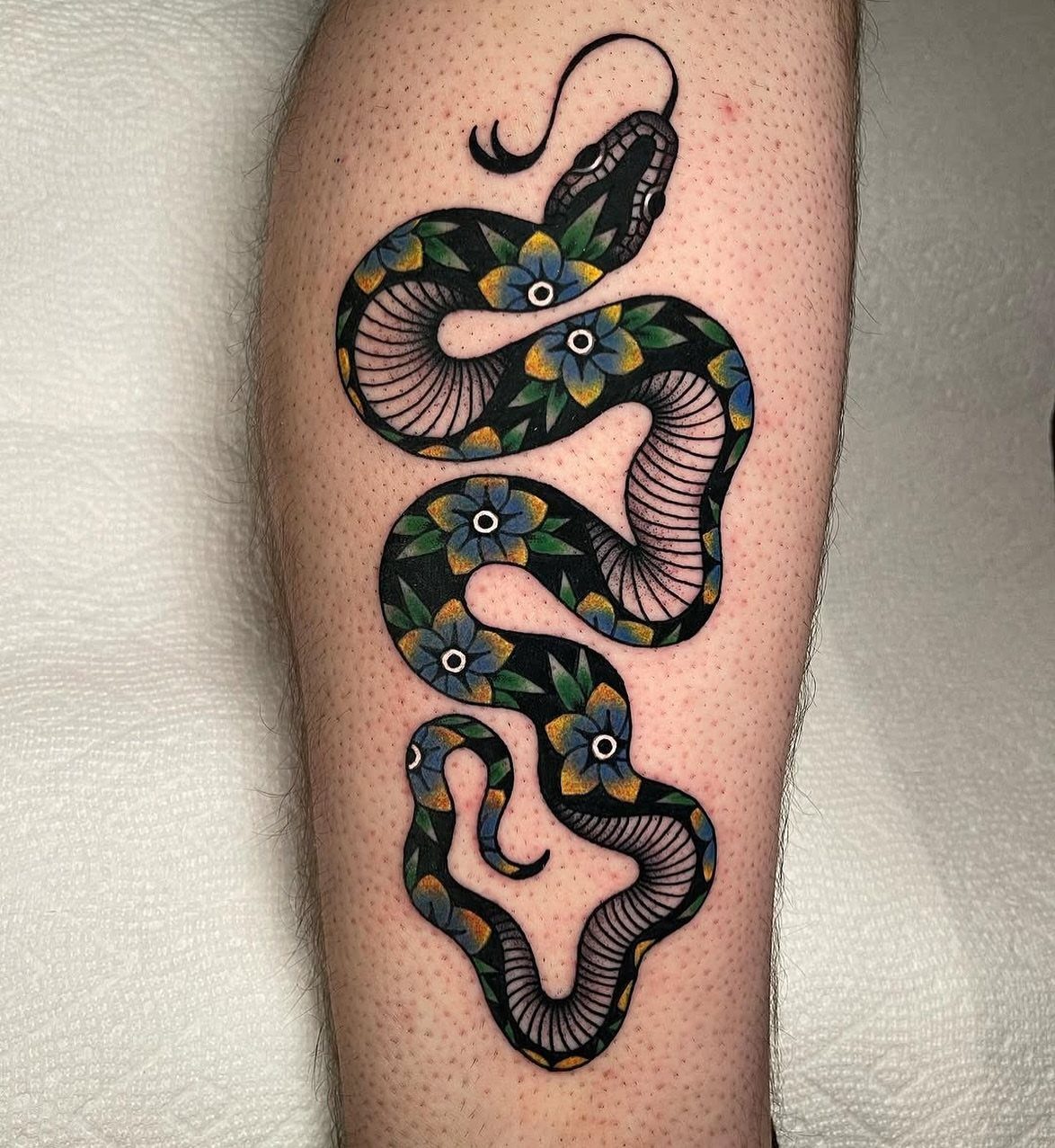 A traditional black snake tattoo with blue and yellow flowers on a person's leg.